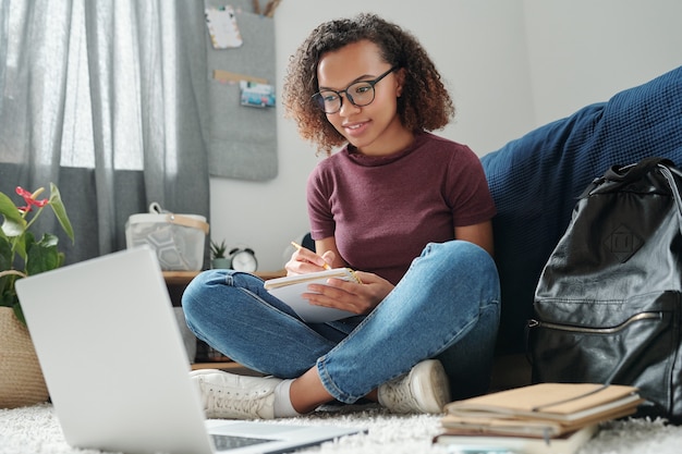 Jeune étudiant multiracial aux jambes croisées en tenue décontractée assis sur le sol près du lit devant un ordinateur portable tout en faisant ses devoirs dans la chambre