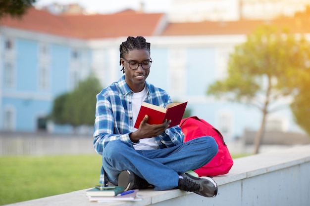 Un jeune étudiant noir heureux en lunettes lisant un livre à l'extérieur.