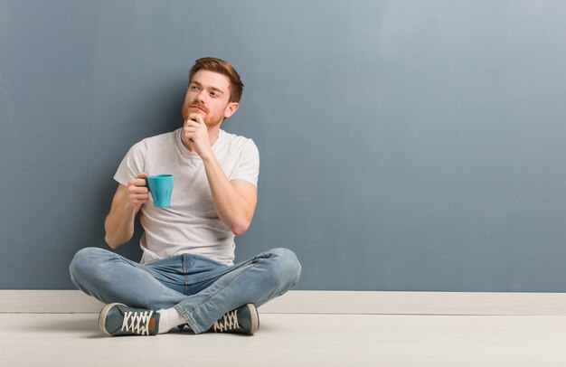 Jeune étudiant rousse homme assis sur le sol doutant et confus. Il tient une tasse de café.