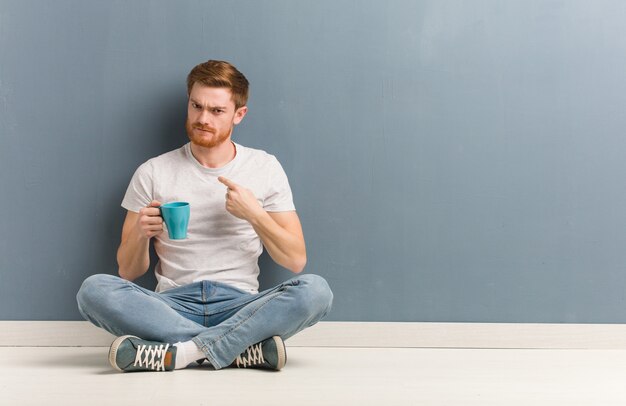 Jeune étudiant rousse homme assis sur le sol invitant à venir. Il tient une tasse de café.