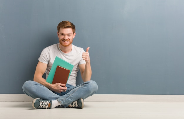 Jeune étudiant rousse homme assis sur le sol en souriant et en levant le pouce. Il tient des livres.