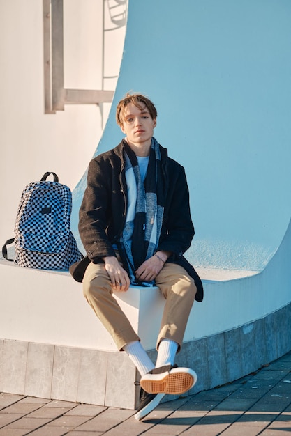 Jeune étudiant avec sac à dos en vieux manteau de laine, écharpe et pantalon court se trouve à l'extérieur