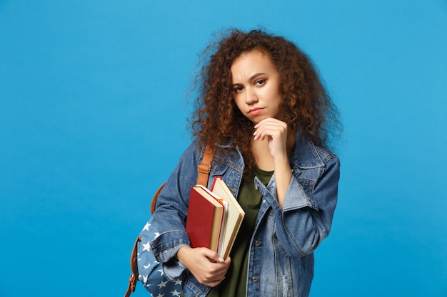 Jeune étudiant triste en vêtements en jean et sac à dos tient des livres isolés sur un mur bleu