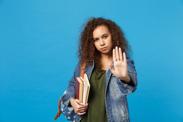 Jeune étudiant triste en vêtements en jean et sac à dos tient des livres isolés sur un mur bleu