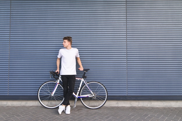 Jeune étudiant avec un vélo blanc se dresse contre le mur et regarde de côté