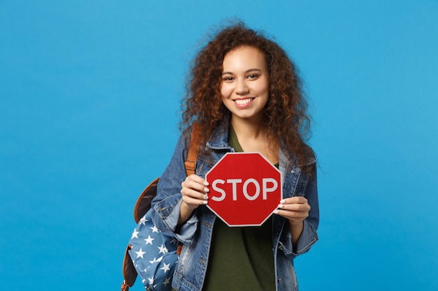 Jeune étudiante adolescente afro-américaine en vêtements en jean, arrêt de prise de sac à dos isolé sur mur bleu