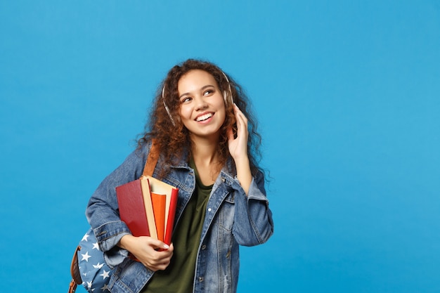 Jeune étudiante adolescente afro-américaine en vêtements en jean, casque d'écoute à dos isolé sur mur bleu