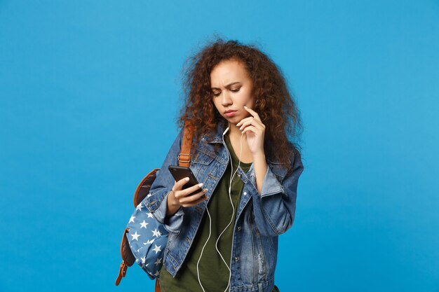 Jeune étudiante adolescente afro-américaine en vêtements en jean, casque d'écoute à dos isolé sur mur bleu