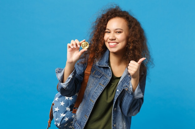 Jeune étudiante adolescente afro-américaine en vêtements en jean, sac à dos tenir bitcoin isolé sur mur bleu