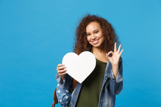 Jeune étudiante adolescente afro-américaine en vêtements en jean, sac à dos tenir coeur isolé sur mur bleu