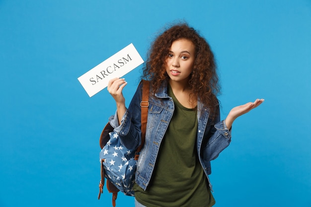 Jeune étudiante adolescente afro-américaine en vêtements en jean, signe de prise de sac à dos isolé sur le mur bleu