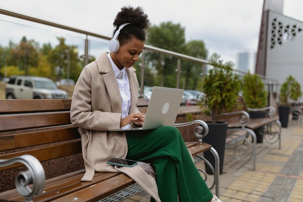 Jeune étudiante afro-américaine travaillant en ligne sur un ordinateur portable avec un casque assis sur un banc