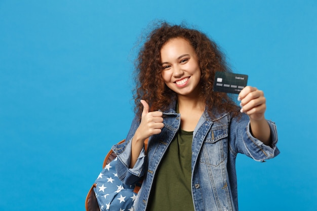 Jeune étudiante afro-américaine en vêtements en jean, sac à dos avec carte de crédit isolée sur mur bleu