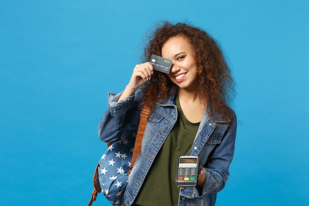 Jeune étudiante afro-américaine en vêtements en jean, sac à dos avec carte de crédit isolée sur mur bleu