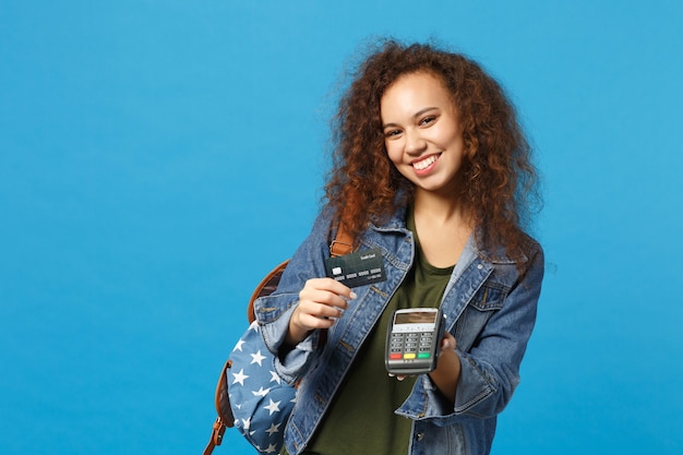 Jeune étudiante afro-américaine en vêtements en jean, sac à dos avec carte de crédit isolée sur mur bleu