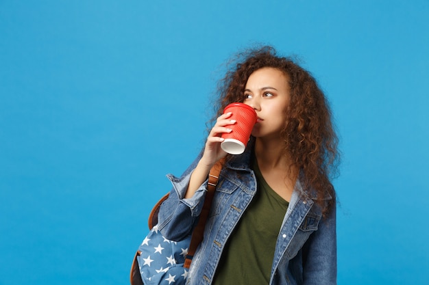 Jeune étudiante afro-américaine en vêtements en jean, sac à dos tenir une tasse en papier isolée sur un mur bleu