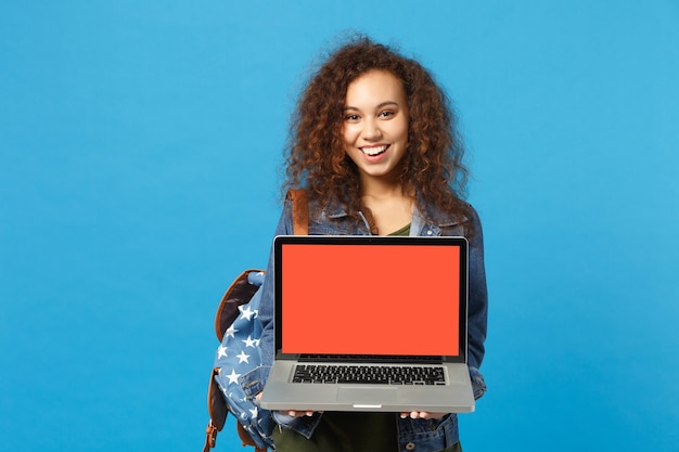 Jeune étudiante afro-américaine en vêtements en jean, travail de sac à dos sur pc isolé sur mur bleu