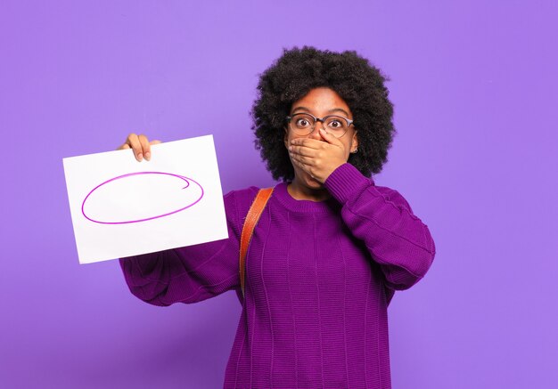 Jeune étudiante afro couvrant la bouche avec les mains avec une expression choquée et surprise, gardant un secret ou disant oups