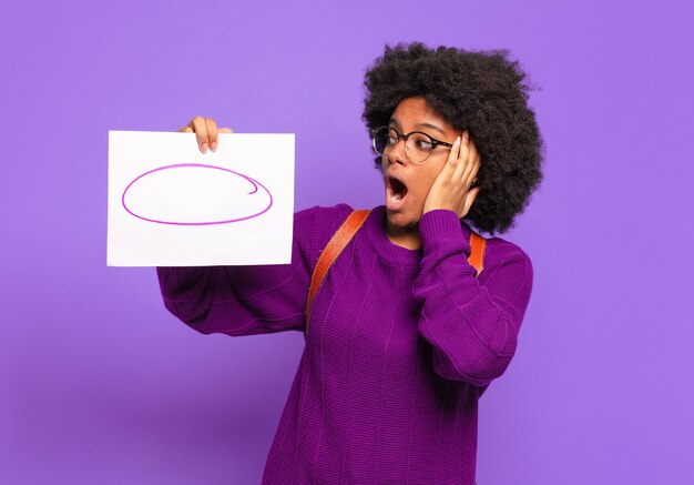 Jeune étudiante afro se sentir heureuse, excitée et surprise, regardant sur le côté avec les deux mains sur le visage