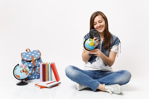 Jeune étudiante agréable et séduisante regardant sur le globe à l'aide d'une loupe assise près du sac à dos, livres scolaires isolés sur mur blanc