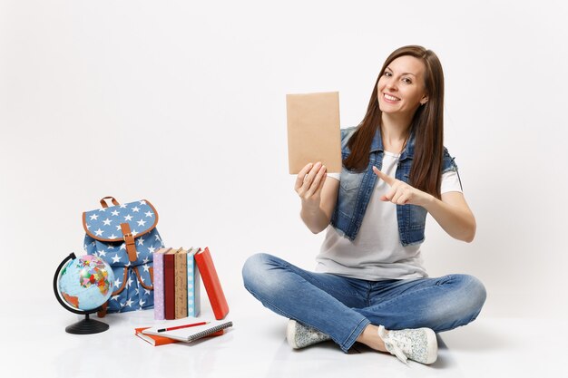 Jeune étudiante agréable en vêtements en denim pointant l'index sur un livre assis près du sac à dos globe, livres scolaires isolés