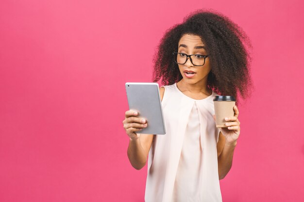 Jeune étudiante américaine aux cheveux bouclés africains tenant une tablette numérique et du café ou du thé sur rose avec espace de copie pour le texte, le logo ou la publicité.