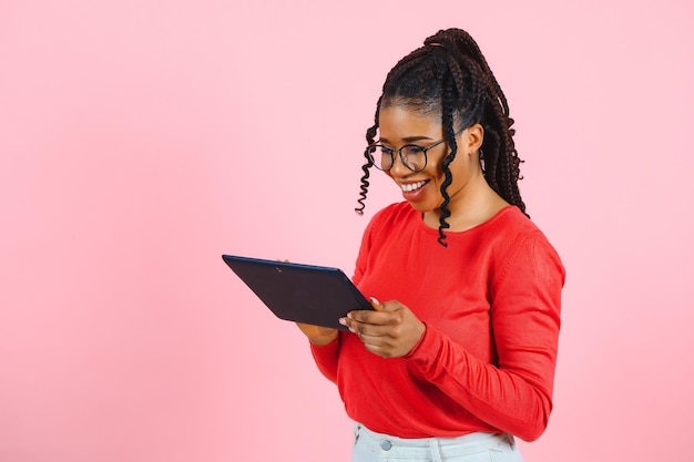 Jeune étudiante américaine aux cheveux bouclés africains tenant une tablette numérique et souriant debout sur fond rose isolé avec espace de copie pour le logo texte ou la publicité