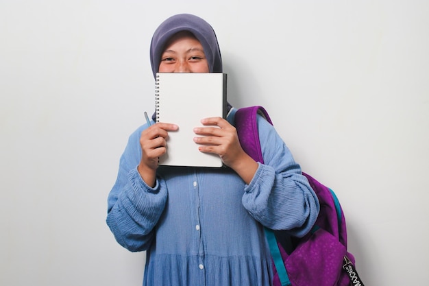 Jeune étudiante asiatique en hijab couvrant son visage avec un livre isolé sur fond blanc