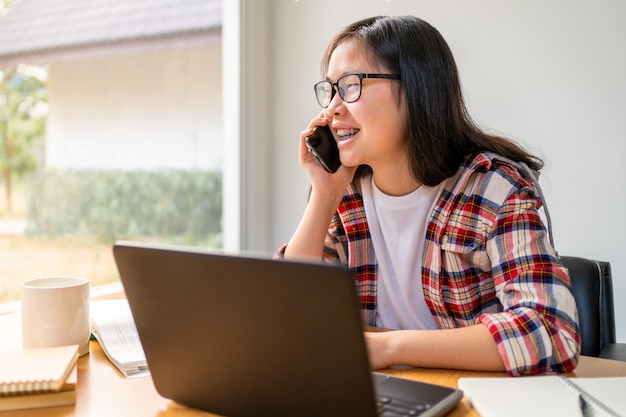 Jeune étudiante asiatique parlant à son amie sur son téléphone tout en travaillant et en étudiant à la maison