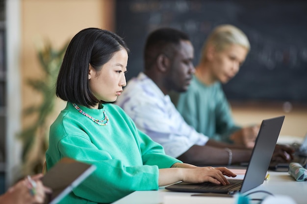 Jeune étudiante asiatique sérieuse en pull vert regardant l'écran d'un ordinateur portable et tapant tout en analysant ou en décodant des données contre ses camarades de classe