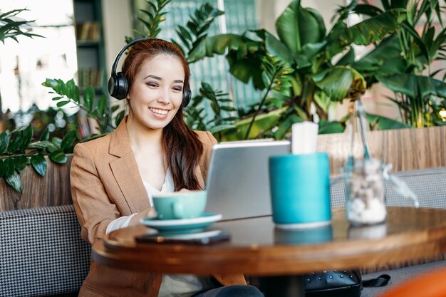 Jeune étudiante asiatique souriante dans les écouteurs communique par tablette apprendre une langue étrangère au café