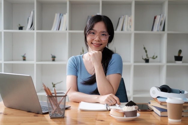 Une jeune étudiante asiatique souriante étudie pour ses cours en ligne avec un ordinateur portable pour se préparer à son examen de mi-semestre.