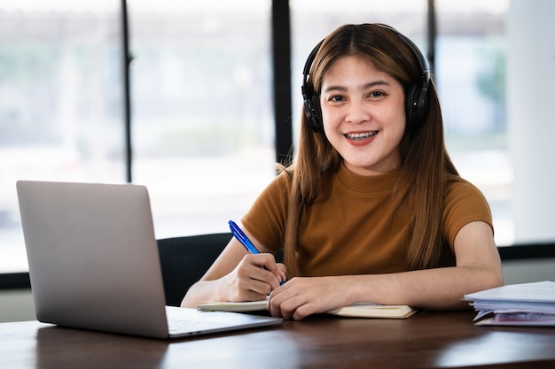 Jeune étudiante asiatique souriante porte des écouteurs sans fil écrivez sur le cahier pour étudier la langue en ligne, regardez et écoutez le conférencier, webinaire par appel vidéo e-learning à la maison, enseignement à distance