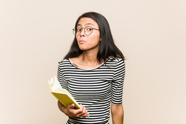 Jeune étudiante asiatique tenant un livre hausse les épaules et les yeux ouverts confus.