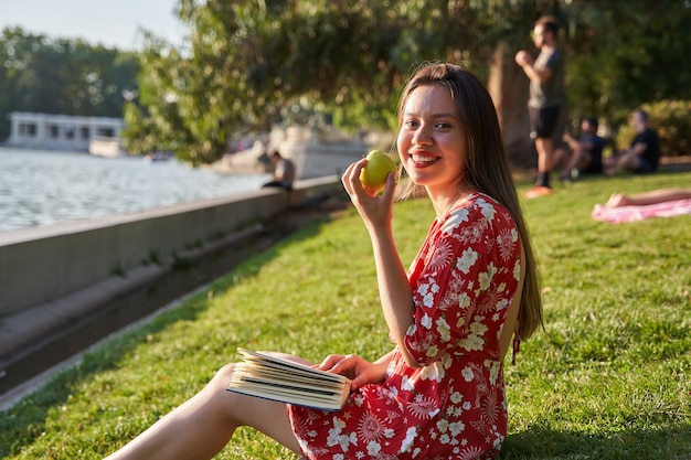 Jeune étudiante assise dans un parc avec un livre en mangeant une pomme