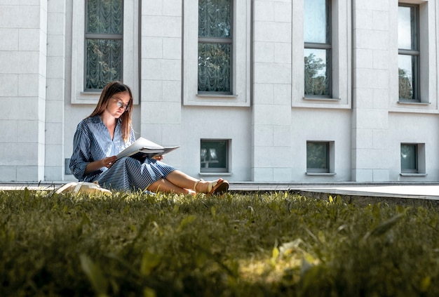 Jeune étudiante assise sur l'herbe près du bâtiment de l'université et apprenant la lecture de manuels jeunes fe...