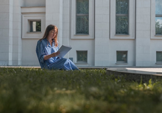 Jeune étudiante assise sur l'herbe près du bâtiment de l'université apprenant et lisant tenant des cours universitaires ...