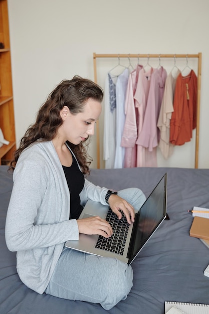 Jeune étudiante assise sur le lit à la maison et travaillant sur un ordinateur portable