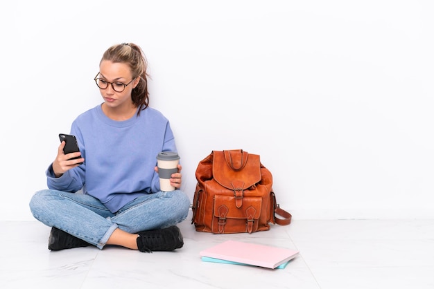Jeune étudiante assise sur le sol isolé sur fond blanc tenant un café à emporter et un mobile