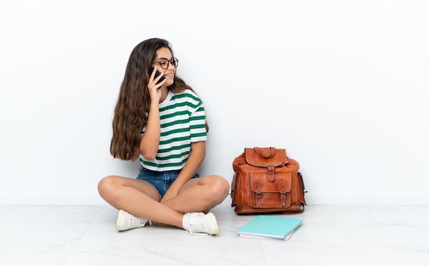 Jeune étudiante assise sur le sol isolée sur fond blanc en gardant une conversation avec le téléphone portable avec quelqu'un