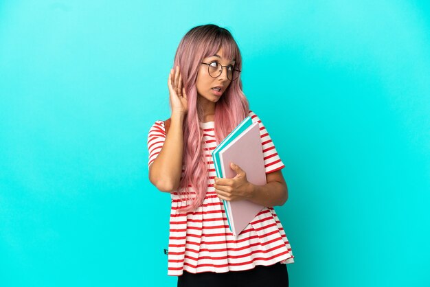Jeune étudiante aux cheveux roses isolée sur fond bleu écoutant quelque chose en mettant la main sur l'oreille