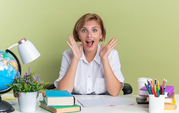 Jeune étudiante blonde excitée assise au bureau avec des outils scolaires regardant la caméra montrant des mains vides isolées sur un mur vert olive