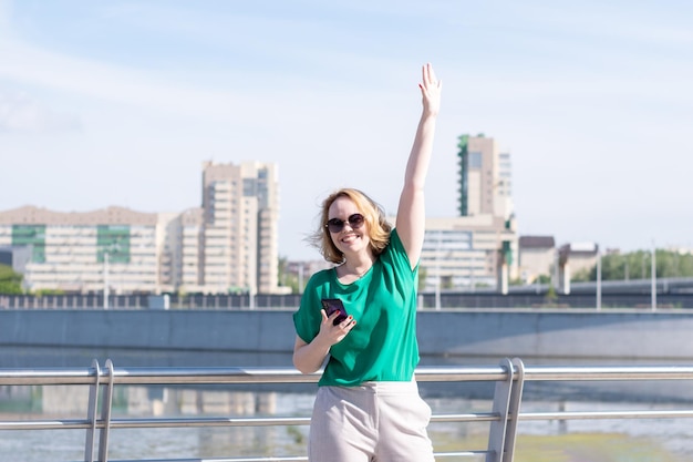 Une jeune étudiante blonde heureuse en lunettes de soleil avec un téléphone en agitant la main en regardant la caméra sur le fond des immeubles de bureaux rivière et ciel bleu La fille accueille des amis Date