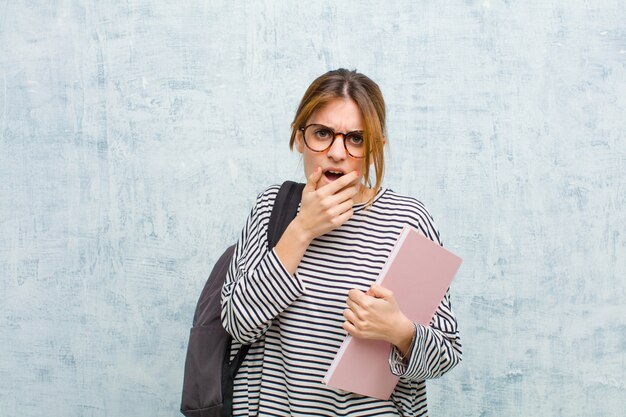 Jeune étudiante avec la bouche et les yeux grands ouverts et la main sur le menton, se sentant désagréablement choquée, disant un mur de grunge whatwow