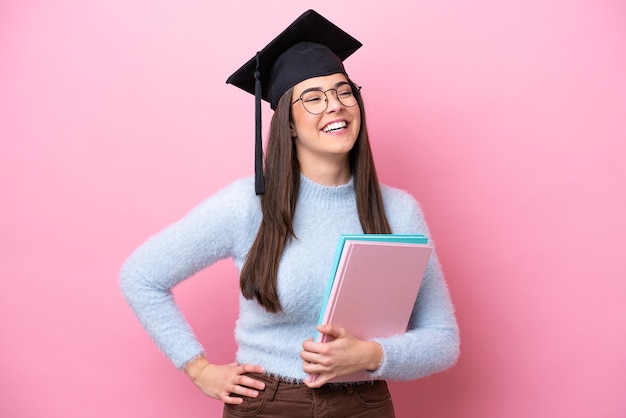 Jeune étudiante brésilienne portant un chapeau diplômé isolé sur fond rose posant avec les bras à la hanche et souriant