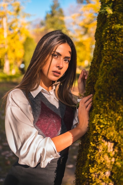 Une jeune étudiante brune de race blanche, dans un parc à côté d'un arbre un après-midi d'automne dans une pose à la mode