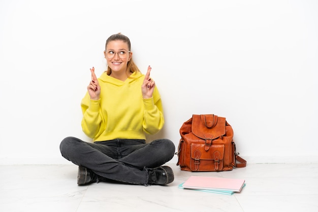 Jeune étudiante caucasienne femme assise sur le sol isolé sur fond blanc avec les doigts croisés