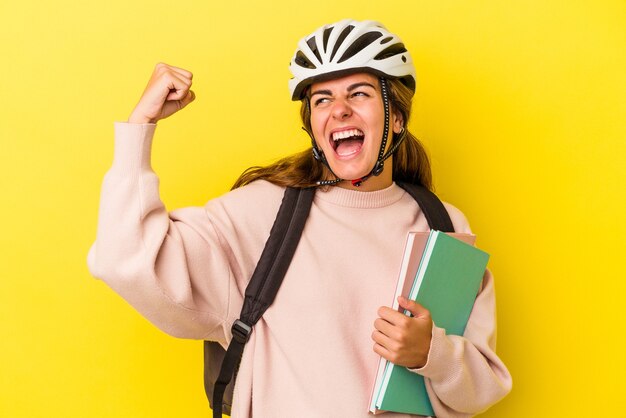 Jeune étudiante caucasienne portant un casque de vélo isolé sur fond jaune levant le poing après une victoire, concept gagnant.