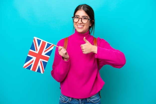 Jeune étudiante caucasienne tenant un drapeau anglais isolé sur fond bleu donnant un geste du pouce levé