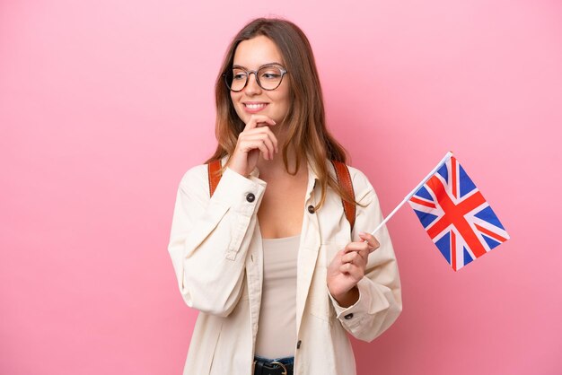 Jeune étudiante caucasienne tenant un drapeau du Royaume-Uni isolé sur fond rose regardant sur le côté et souriant